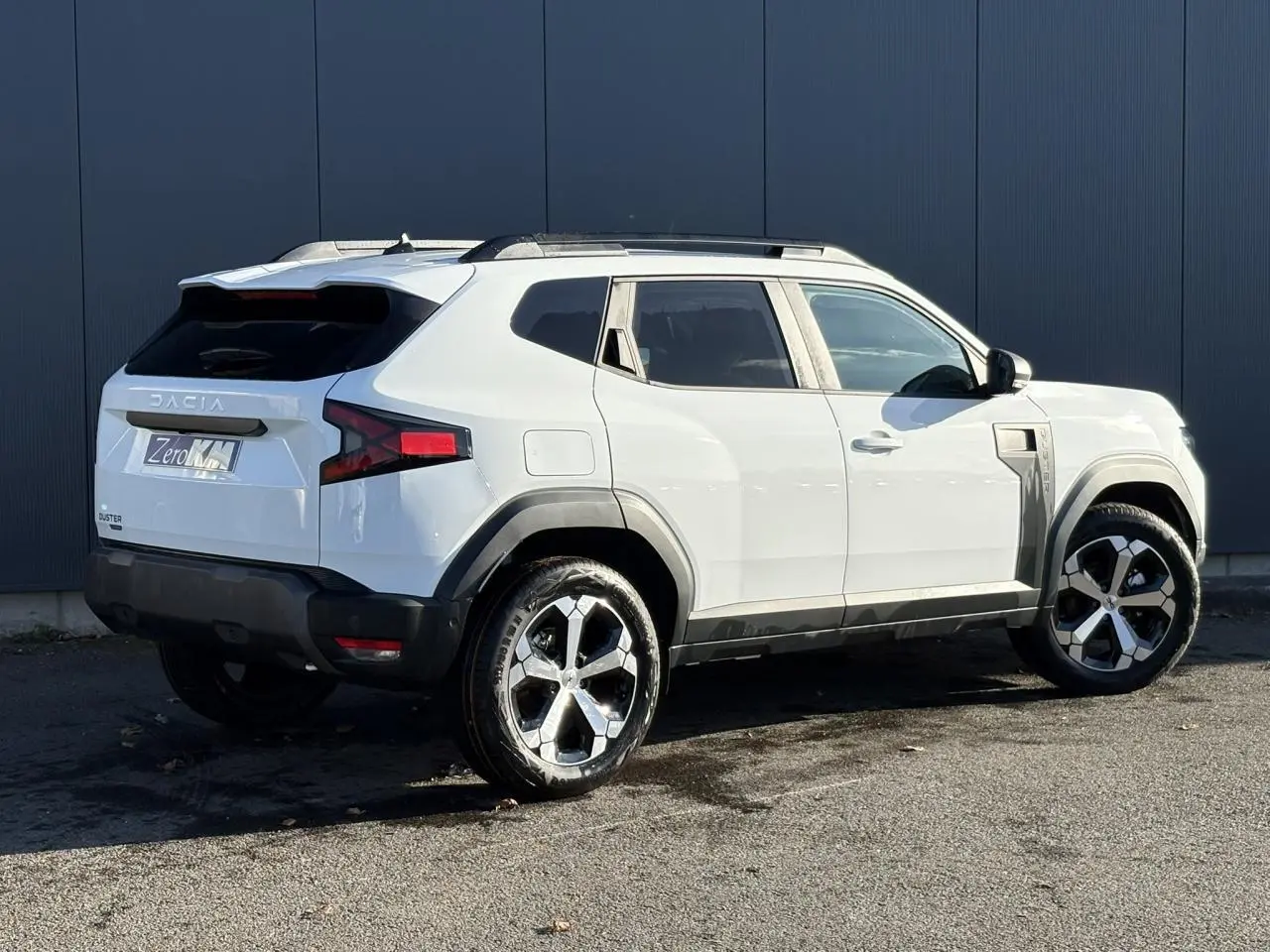 Vue 3/4 arrière droite du Dacia Duster blanc Glacier avec jantes alliage et barres de toit noires.
