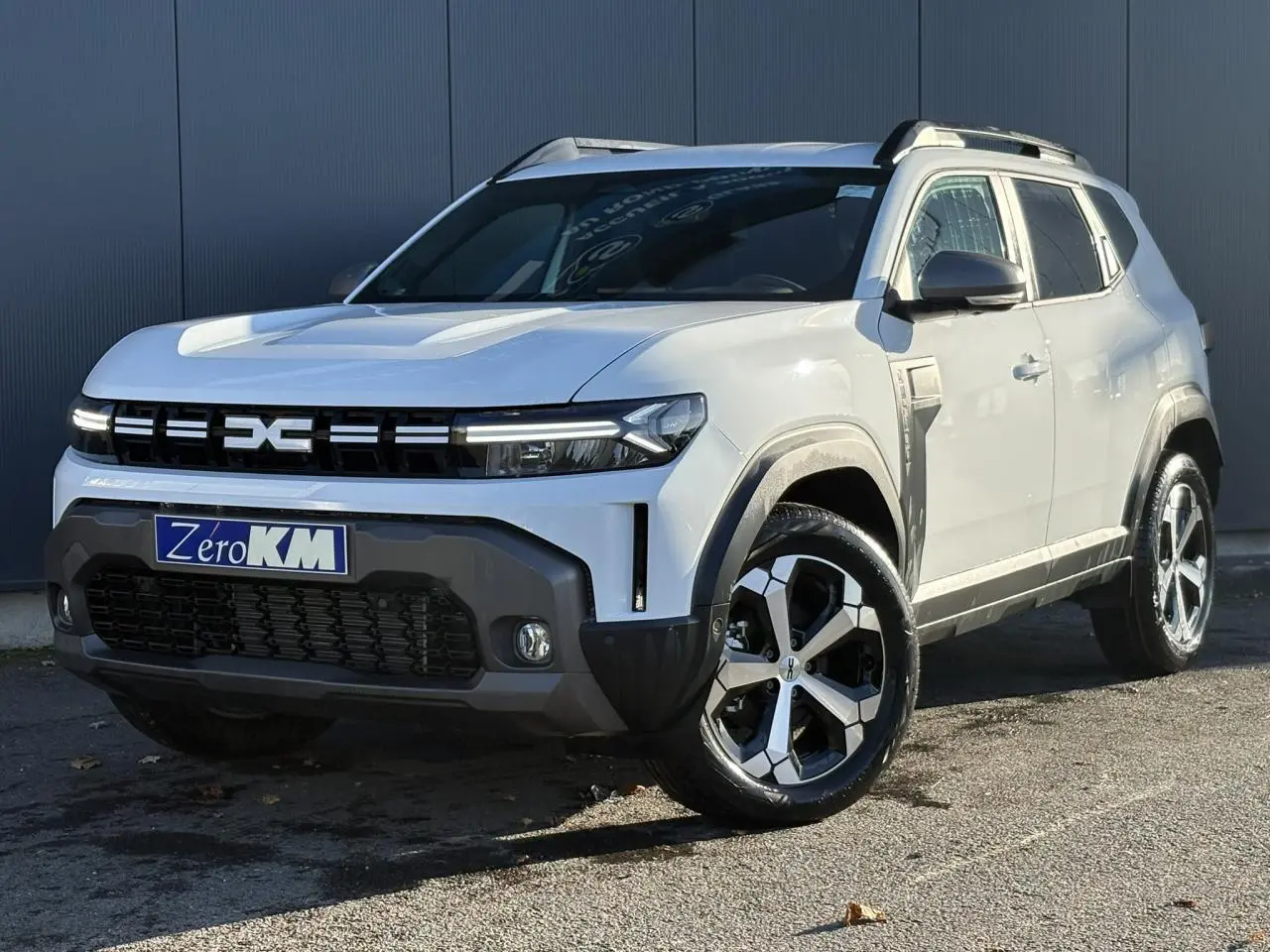 Dacia Duster blanc glacier en 3/4 avant droit, avec jantes alliage noires et grises et barres de toit noires.