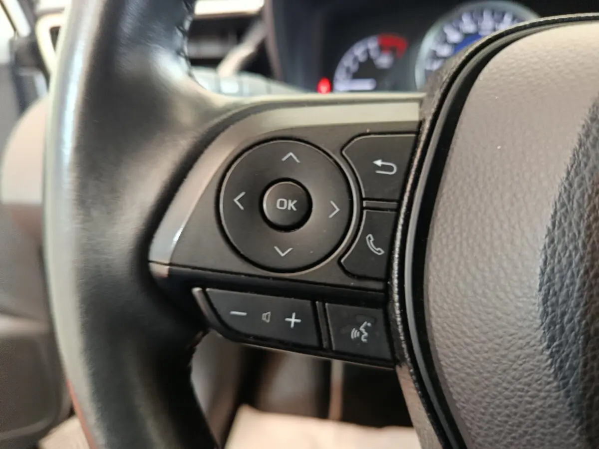 Gros plan sur les commandes gauche du volant cuir noir de la Toyota Corolla Hybride 122h Active, avec tableau de bord flou en arrière-plan.