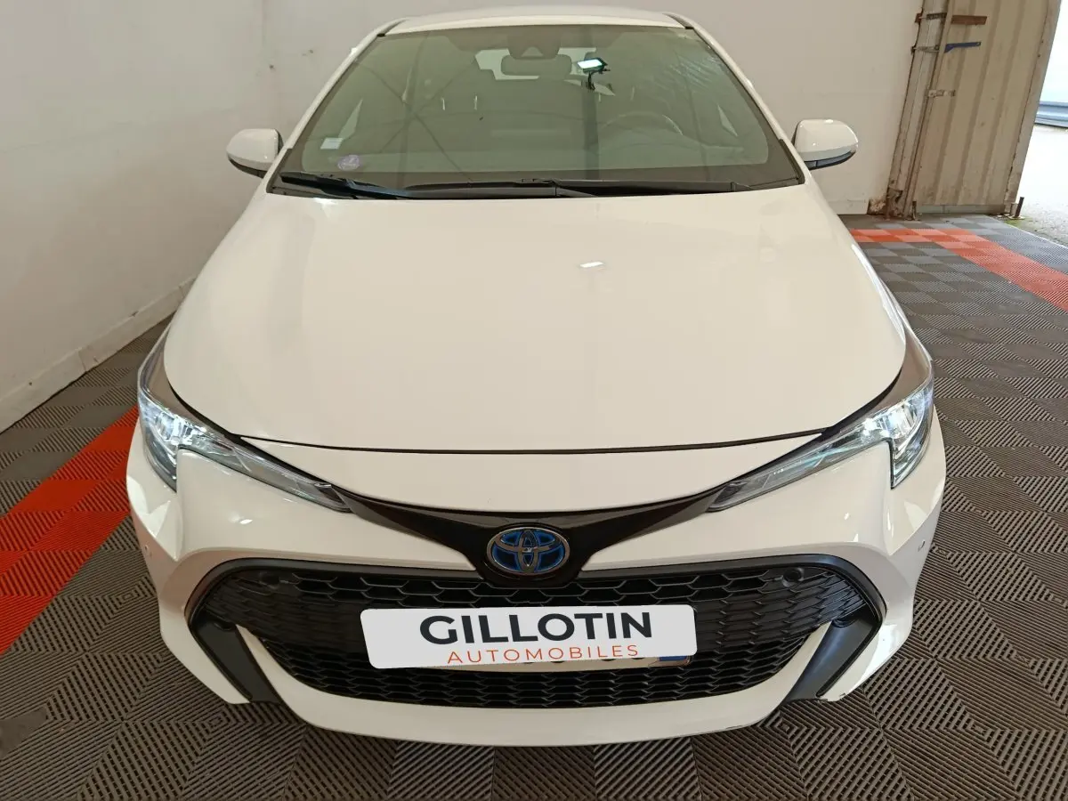 Vue avant d'une Toyota Corolla Hybride 122h Active blanche avec calandre noire et logo bleu Toyota visible.