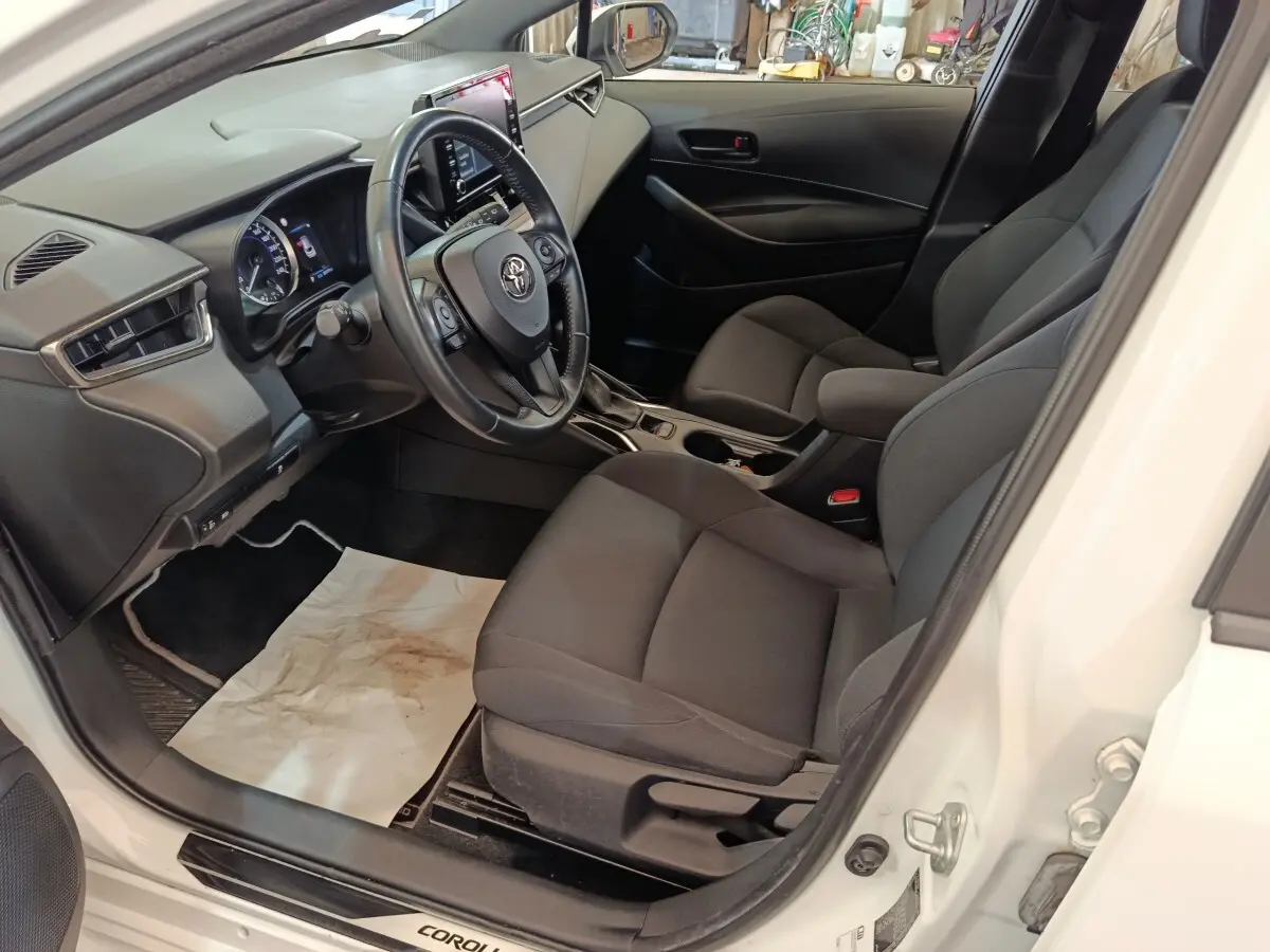 Vue intérieure côté conducteur de la Toyota Corolla Hybride blanche 2020, avec tableau de bord et sièges noirs.