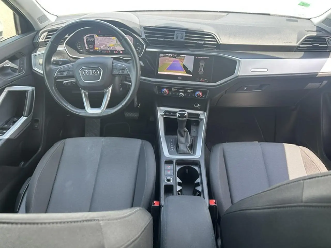 Vue intérieure avant de l'Audi Q3 blanc, montrant le volant multifonctions, écran central avec caméra de recul et sellerie tissu noire.