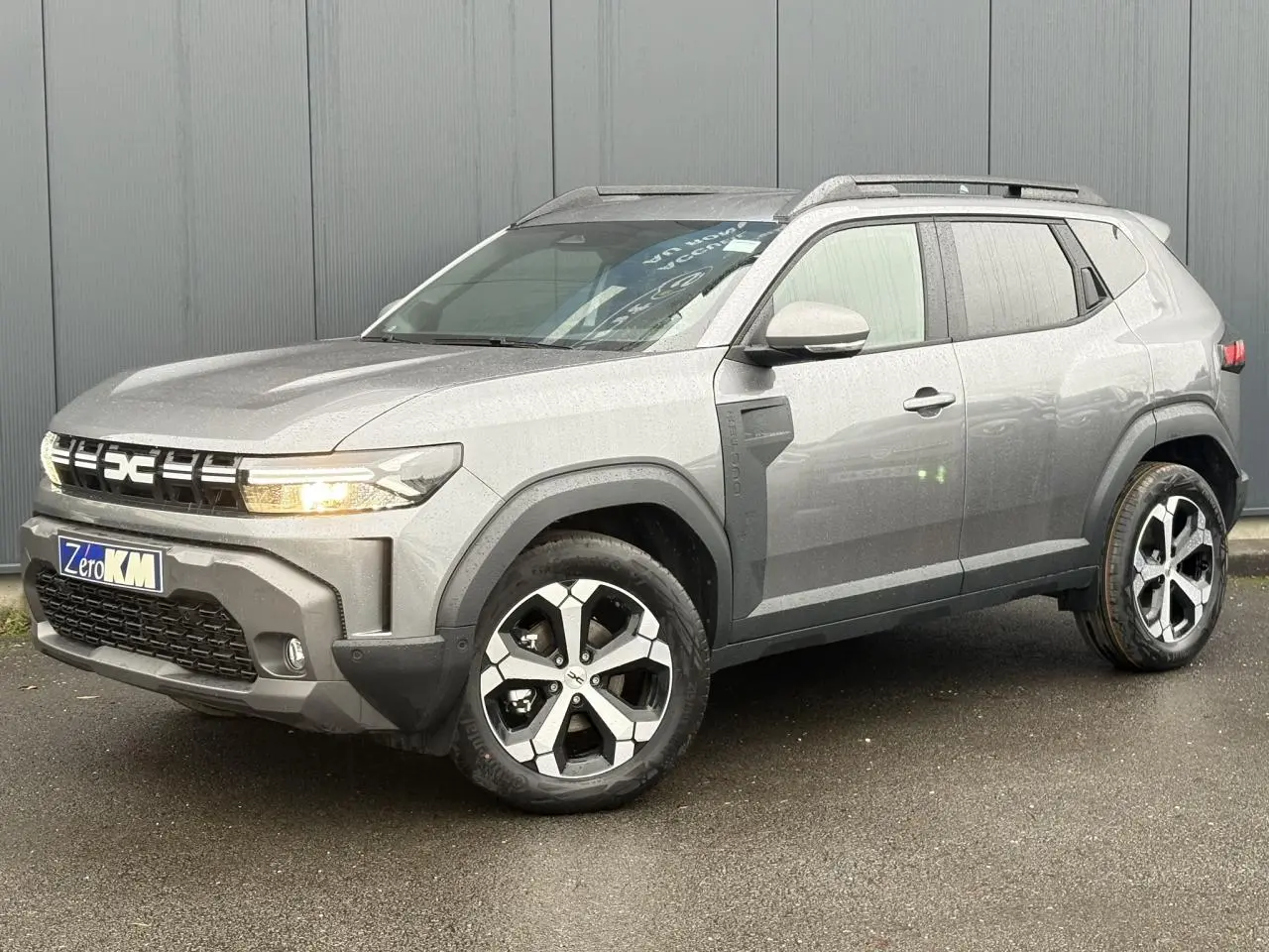 Dacia Duster gris schiste en 3/4 avant droit, avec jantes alliage diamantées et barres de toit noires.