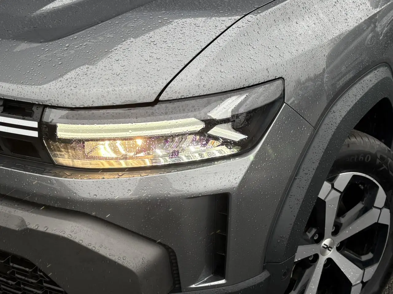 Gros plan sur l'avant droit gris schiste du Dacia Duster 2026, avec phare allumé et jante alliage diamantée.