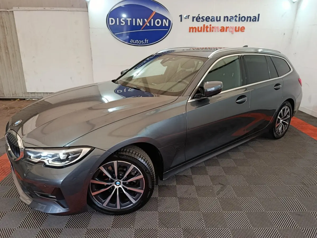 BMW Série 3 318d gris clair vue 3/4 avant droit, avec jantes alliage et phares LED allumés.