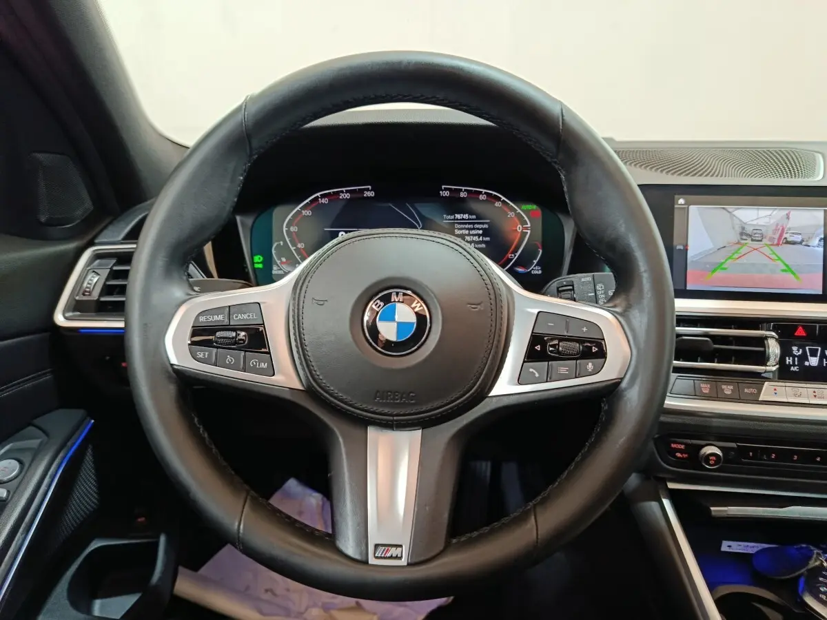 Vue intérieure centrée sur le volant cuir noir BMW avec tableau de bord digital et écran multimédia en arrière-plan.