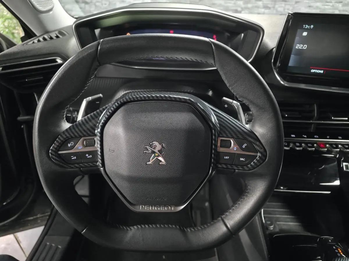 Volant compact cuir noir avec insert carbone et logo Peugeot, vue rapprochée du tableau de bord et écran tactile.