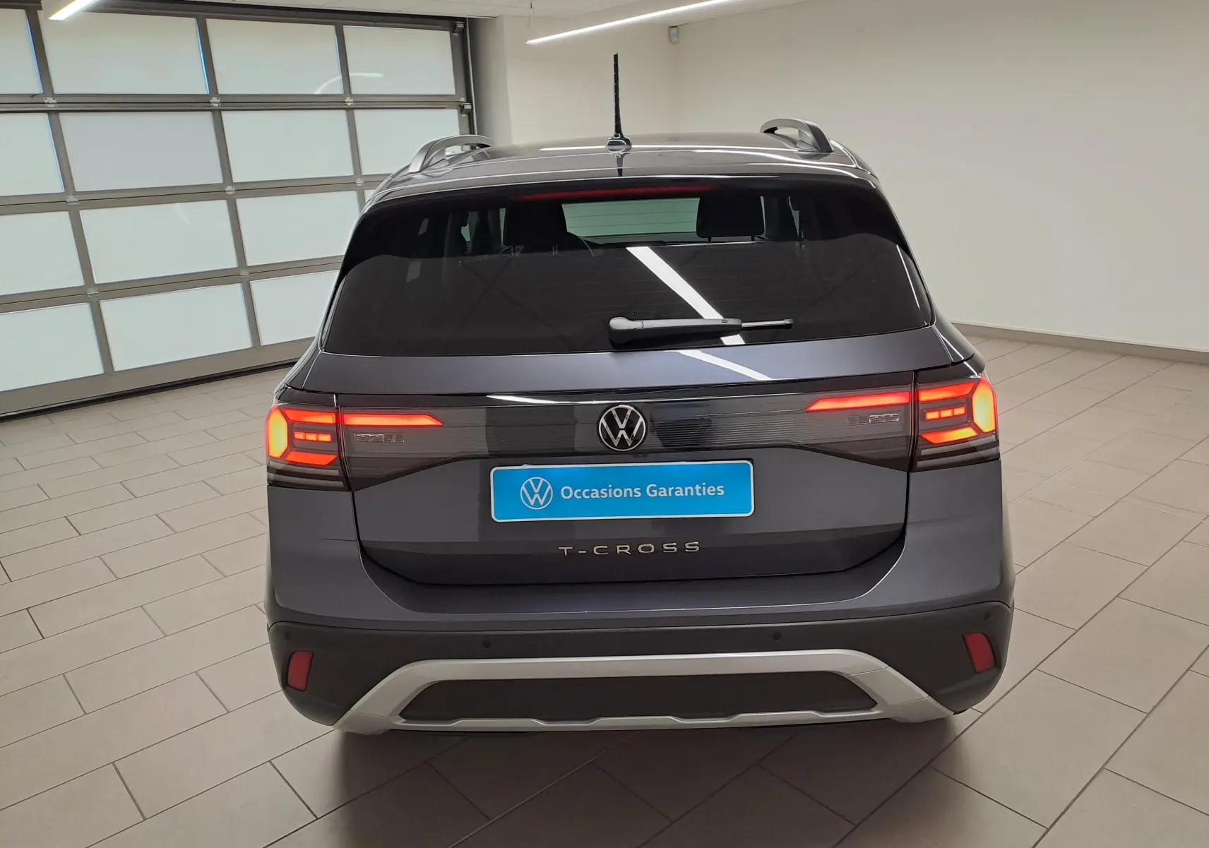 Vue arrière du Volkswagen T-Cross gris cendré 2025 avec feux allumés et logo VW visible en intérieur showroom.