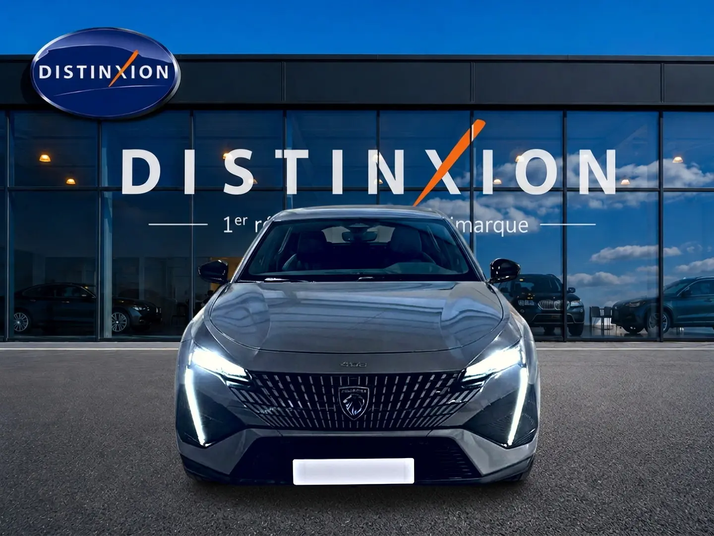 Vue avant d'une Peugeot 408 Hybrid 145 GT gris sélénite avec phares LED allumés devant une vitrine Distinxion.