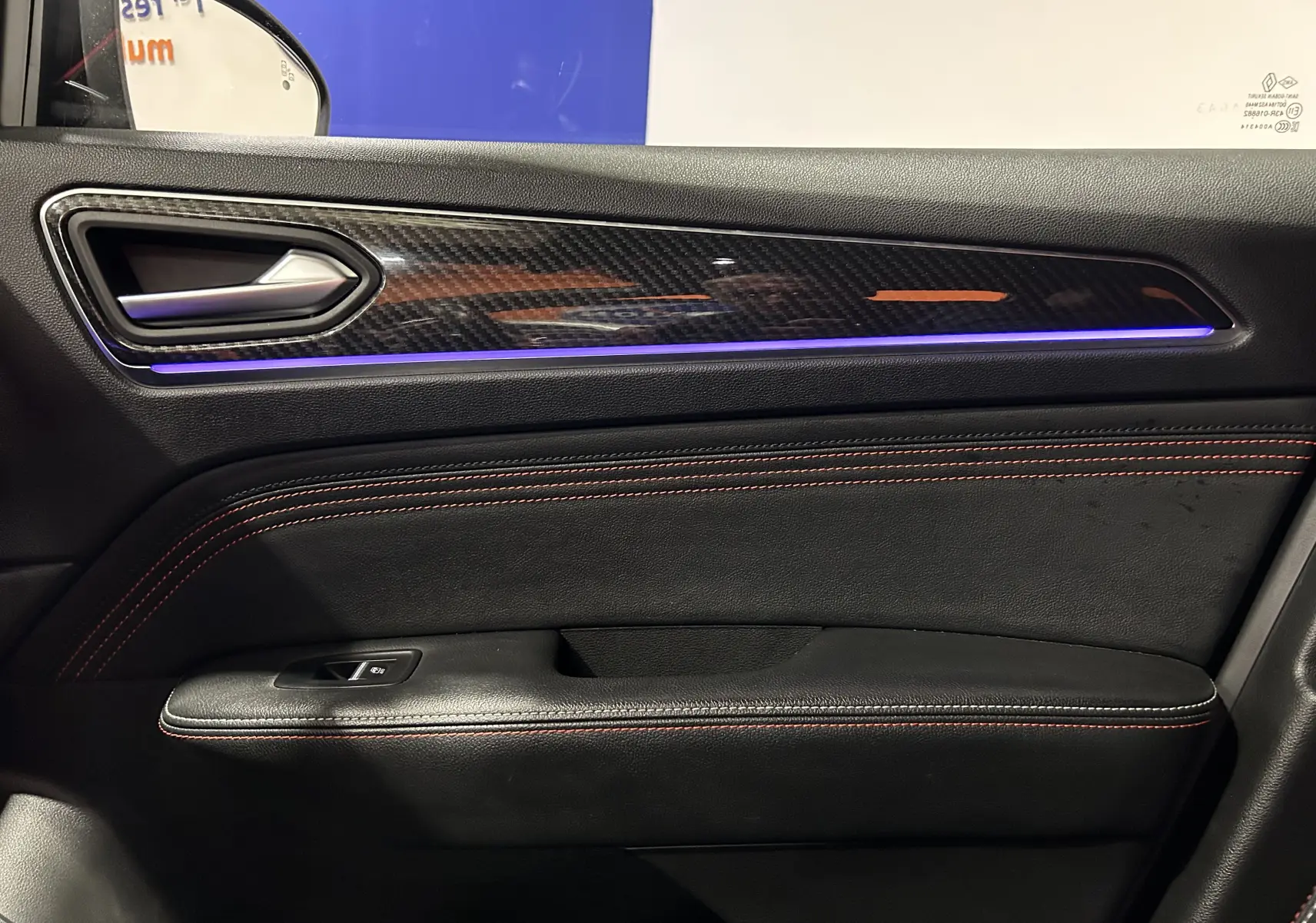 Intérieur côté gauche de porte Renault Arkana noir avec surpiqûres rouges et bandeau décoratif carbone éclairé en bleu.