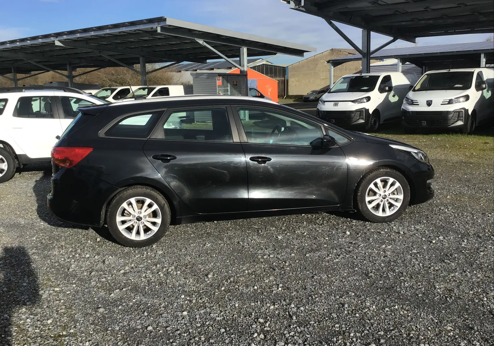 Vue de profil côté gauche d'une Kia CEE'D SW noire de 2017 garée sur un sol gravillonné avec des véhicules utilitaires en arrière-plan.