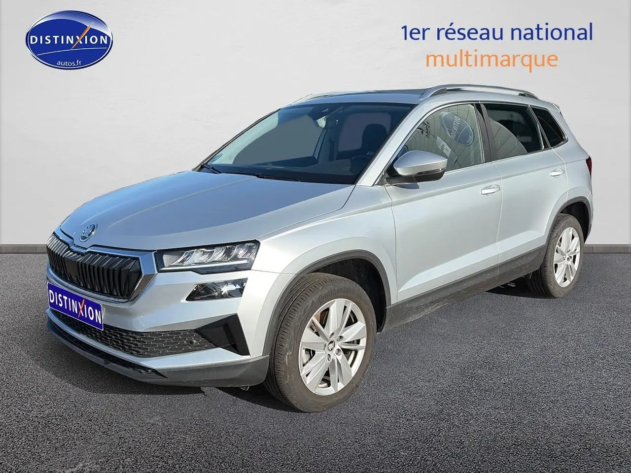 SUV Skoda Karoq gris en 3/4 avant droit avec calandre noire et jantes alu visibles sur sol goudronné.