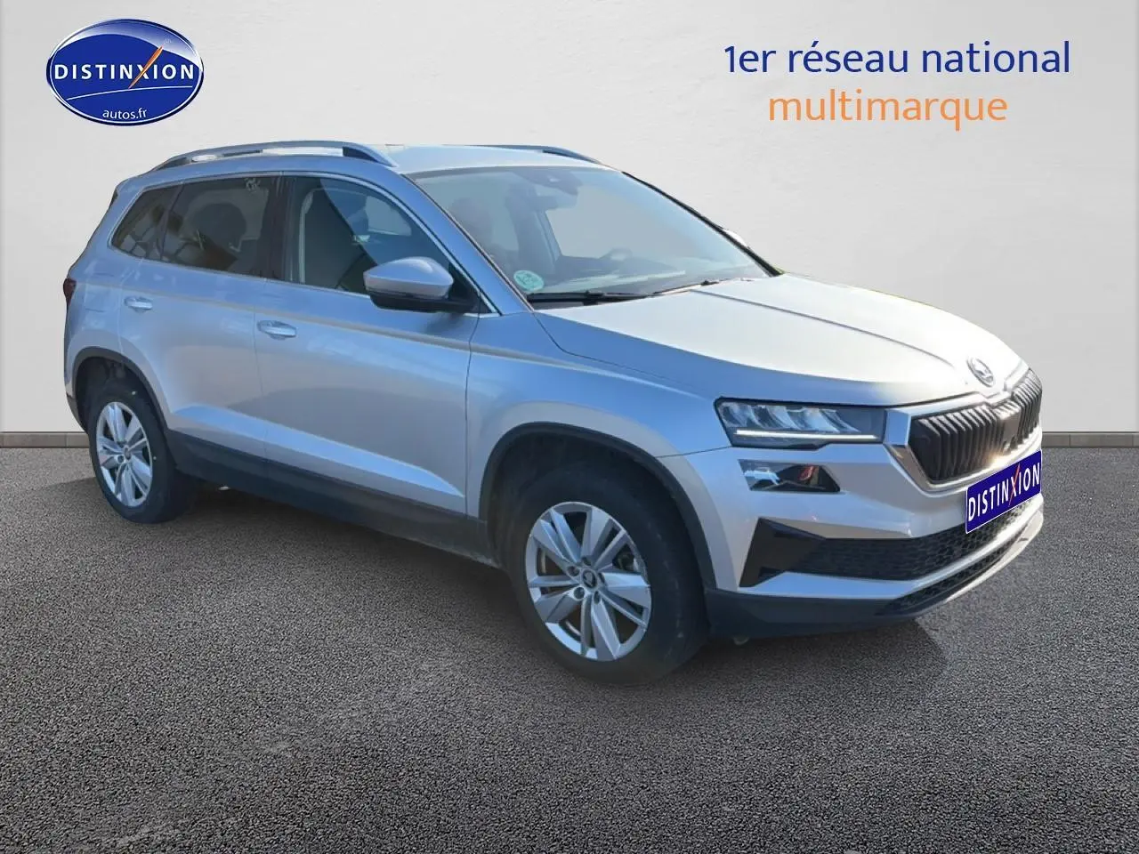 SKODA KAROQ gris 3/4 avant droit, jantes alu, barres de toit et feux LED visibles sur fond neutre.