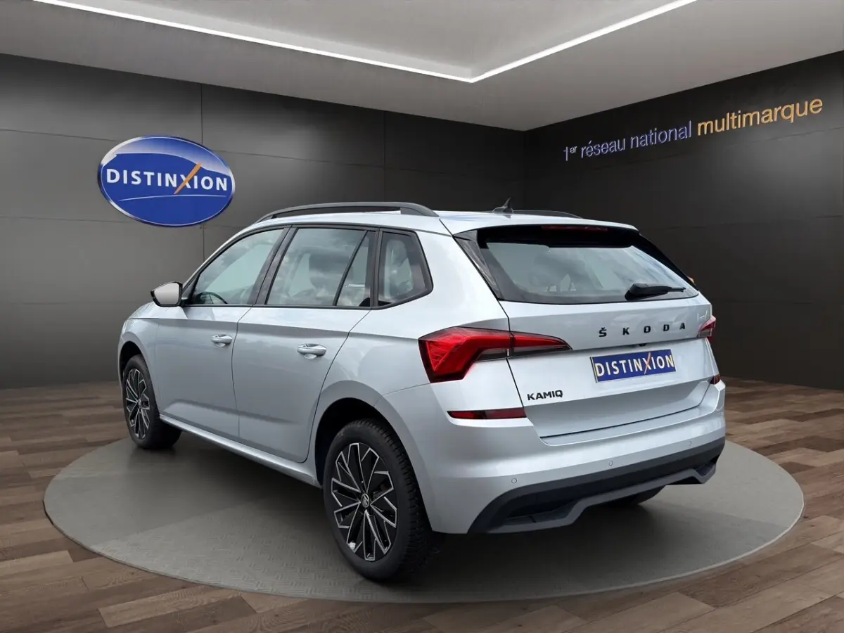 Vue 3/4 arrière droite d'un Skoda Kamiq gris clair avec jantes bi-ton et barres de toit noires en showroom.