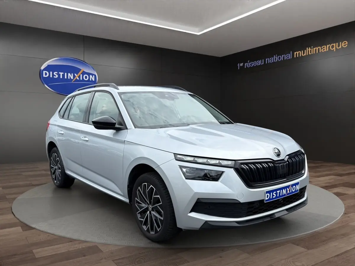 Skoda Kamiq gris clair en 3/4 avant droit, avec jantes bi-ton et rétroviseurs noirs rabattables.