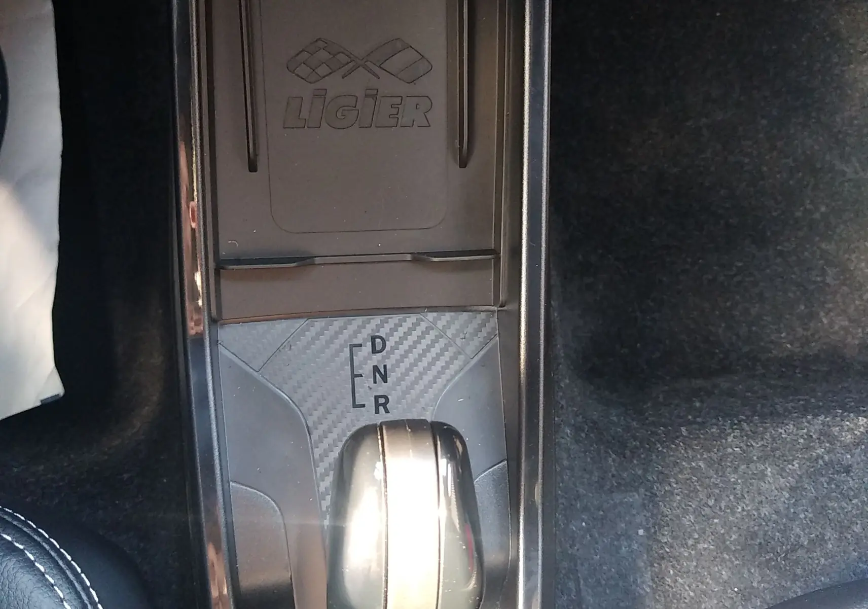 Vue plongeante sur la console centrale noire avec levier de boîte auto et logo Ligier en finition carbone.