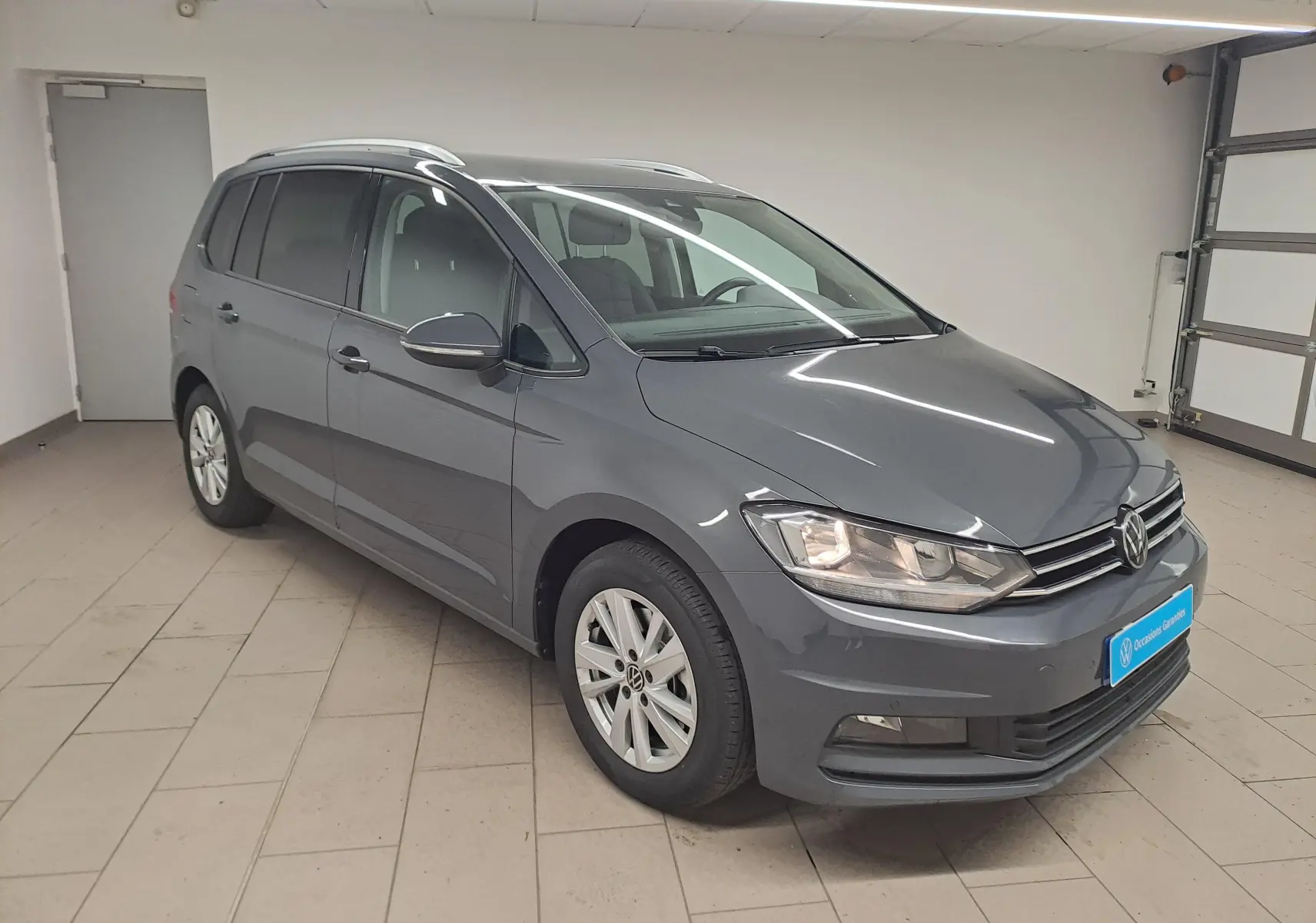 Volkswagen Touran gris dauphin en 3/4 avant droit dans un garage, avec phares allumés et jantes alliage.