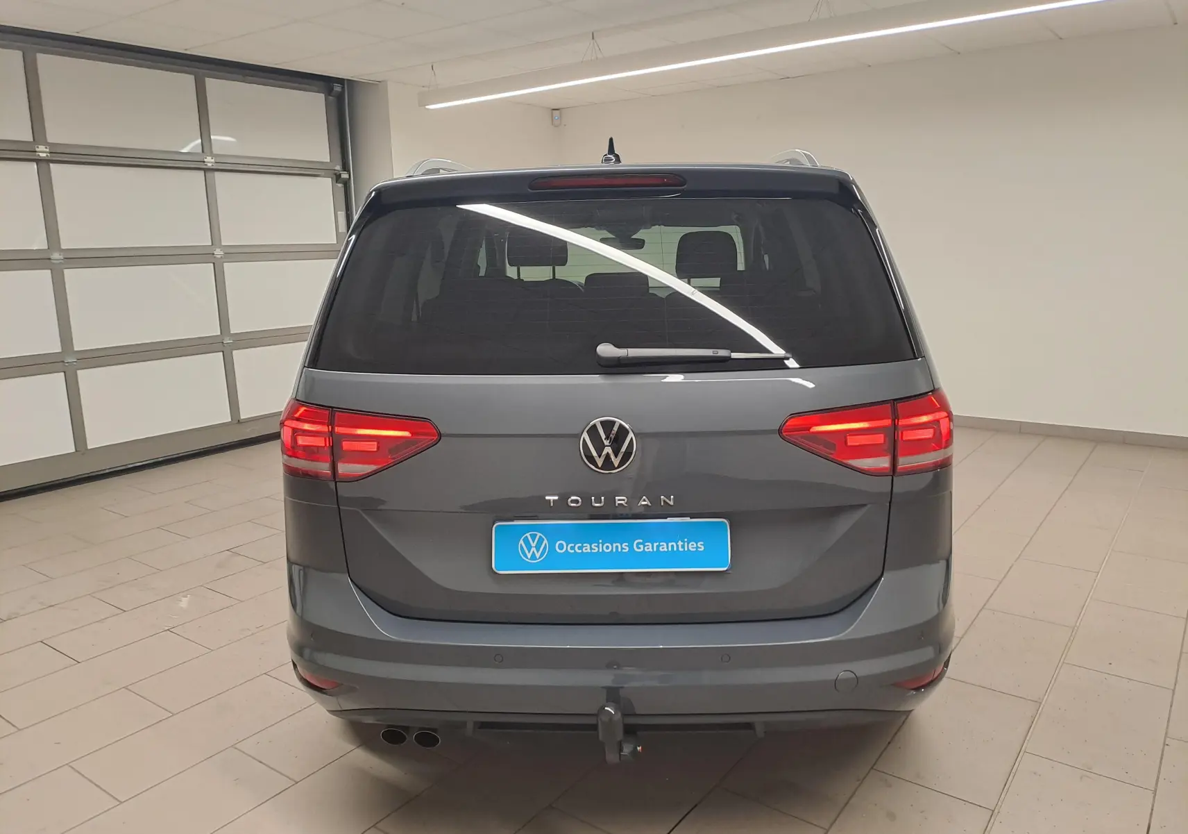 Vue arrière d'un Volkswagen Touran gris dauphin 2024 avec attelage et feux allumés en intérieur.