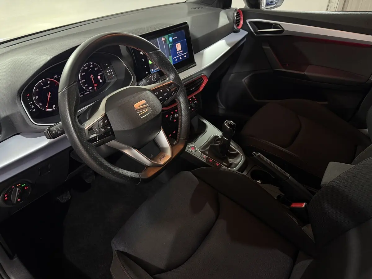 Vue intérieure côté conducteur de la SEAT Ibiza 1.0 TSI 110 FR 2024, volant cuir, écran tactile et détails rouges sur la console.