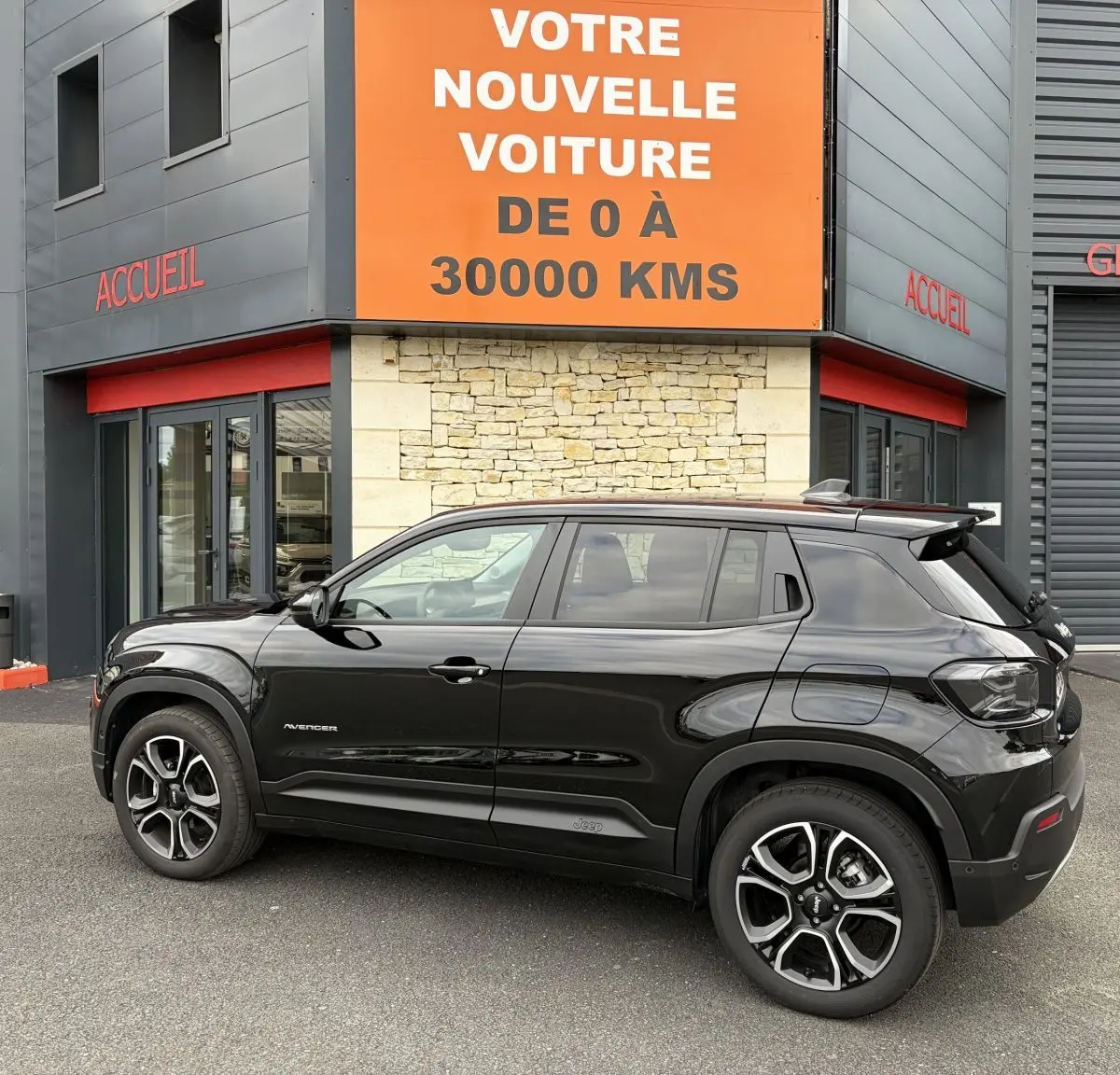 Vue latérale droite d'un Jeep Avenger noir 2025 garé devant un bâtiment avec panneau orange "Votre nouvelle voiture".