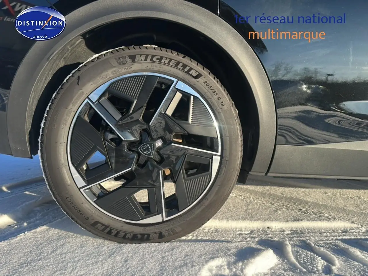 Gros plan sur la roue avant gauche et la jante alu noire du Peugeot 408 Hybrid 145 GT e-dcs6 noir sur sol enneigé.