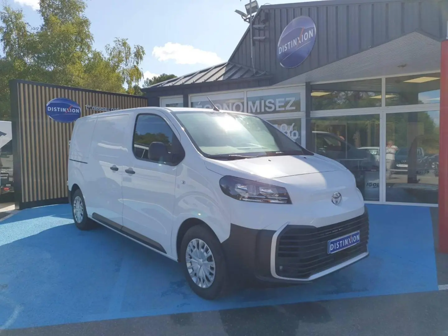 Toyota Proace Fourgon blanc Glacier vu en 3/4 avant droit sur parking bleu devant un bâtiment commercial Distinxion.