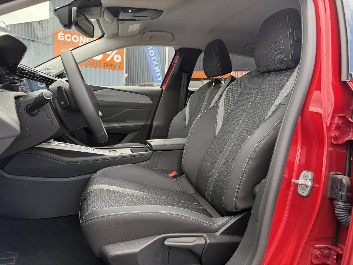 Intérieur côté conducteur d’une Peugeot 308 SW rouge, sièges noirs avec surpiqûres bleues et accoudoir central visible.