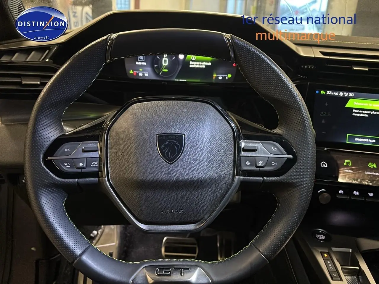 Vue rapprochée du volant cuir noir avec logo Peugeot et commandes intégrées, tableau de bord digital en arrière-plan.