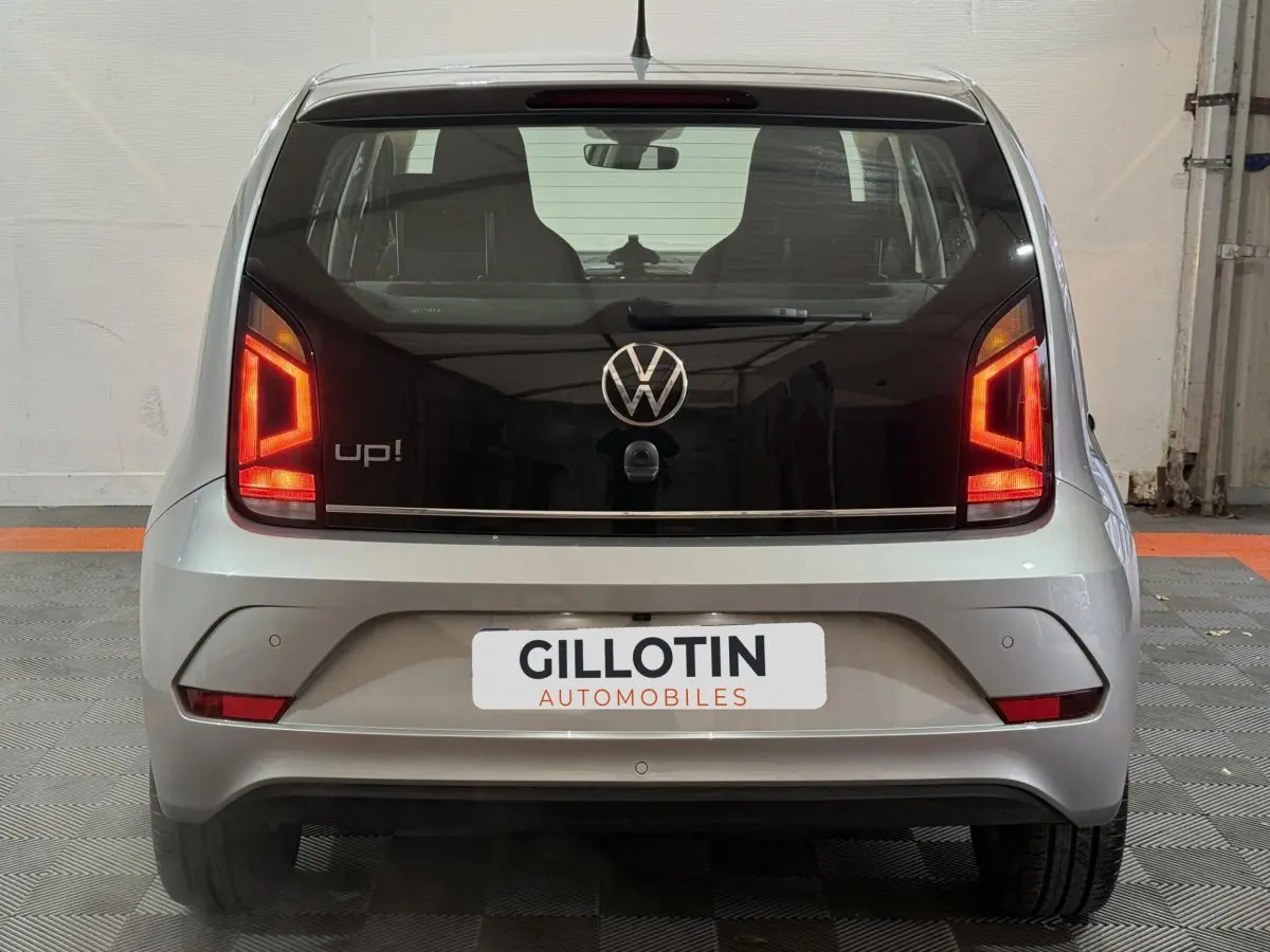 Vue arrière d'une Volkswagen Up! gris foncé avec feux arrière allumés et logo VW visible sur le hayon noir.