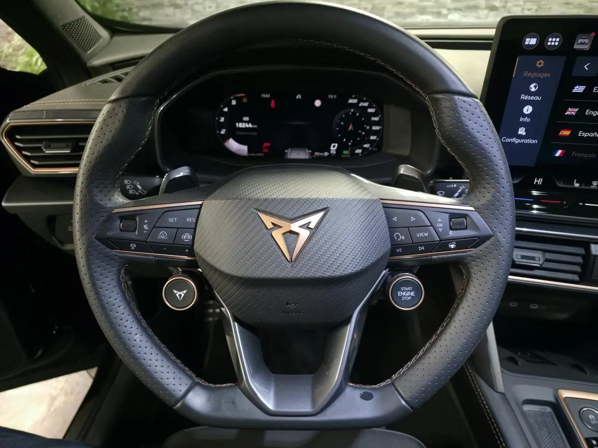 Volant sport noir perforé avec logo CUPRA cuivré, vue frontale du poste de conduite et tableau de bord digital.