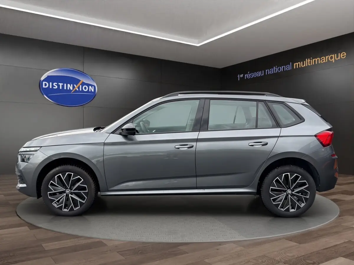 Vue latérale droite d'un Skoda Kamiq gris foncé avec jantes bi-ton et barres de toit noires en showroom.