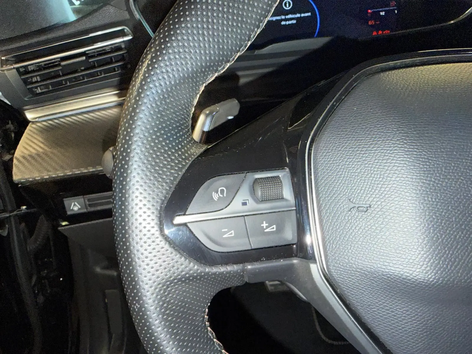 Gros plan sur le volant cuir perforé noir du Peugeot 508 SW 2024 avec commandes intégrées et décor carbone en arrière-plan.