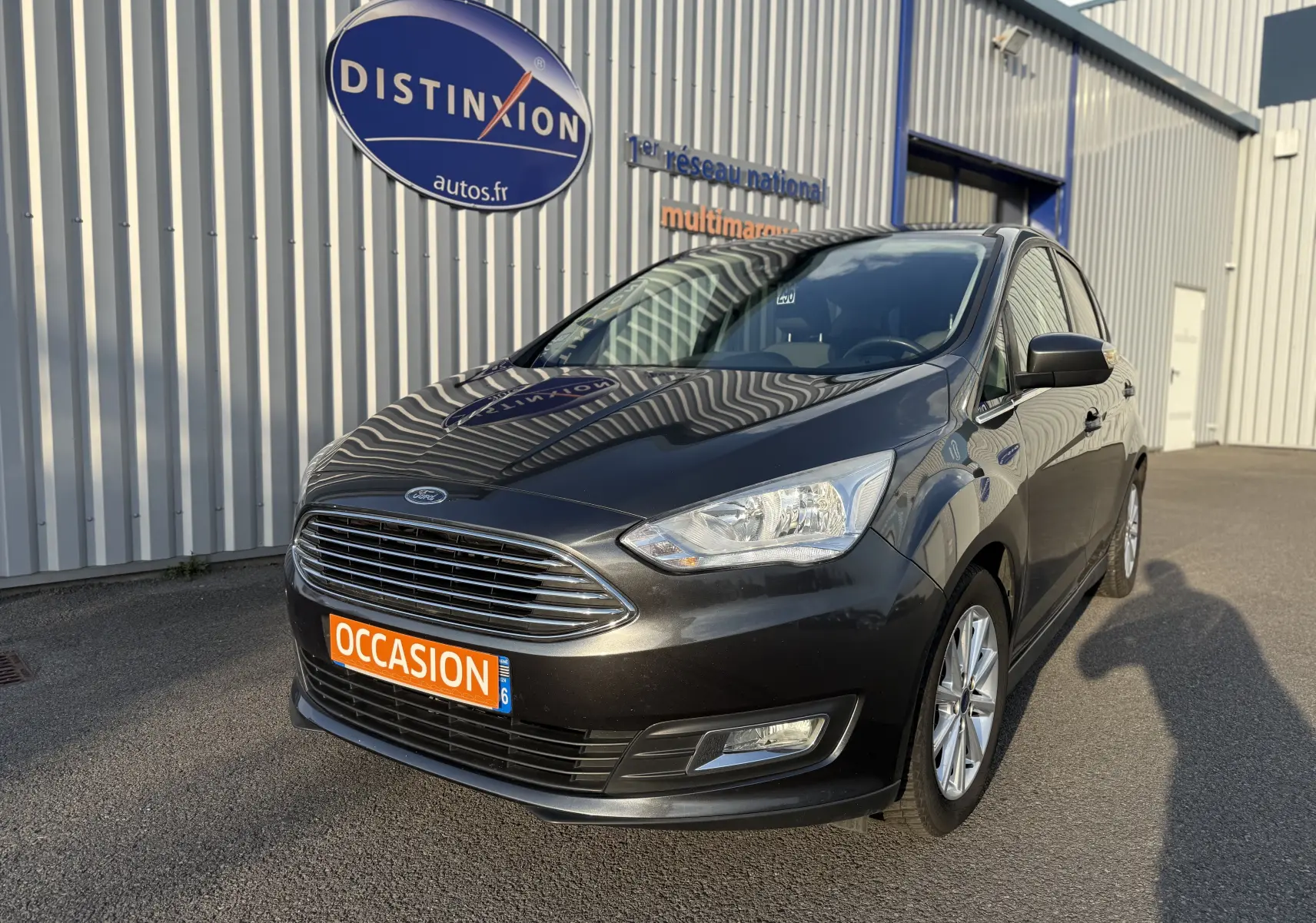 Ford C-MAX 1.6 TDCI 120 Titanium gris foncé vue 3/4 avant avec plaque "OCCASION" devant un bâtiment industriel.
