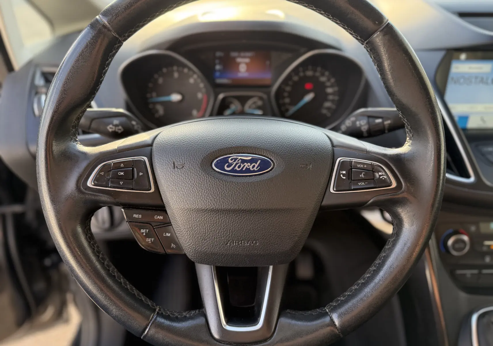Vue rapprochée du volant en cuir noir d’une Ford C-MAX 2018, avec commandes intégrées et tableau de bord flou en arrière-plan.