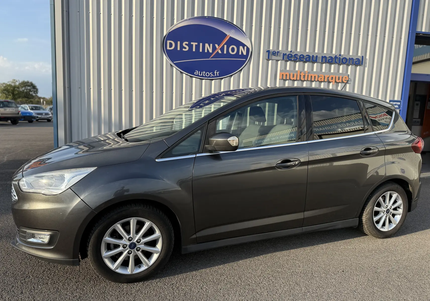 Vue de profil côté gauche d'une Ford C-MAX gris foncé garée devant un bâtiment avec enseigne Distinxion.