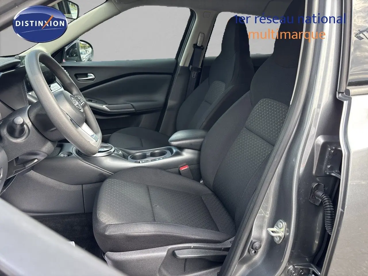 Vue intérieure côté conducteur du Nissan Juke Hybrid 2024 en gris squale métal, sièges tissu noir et volant multifonction.