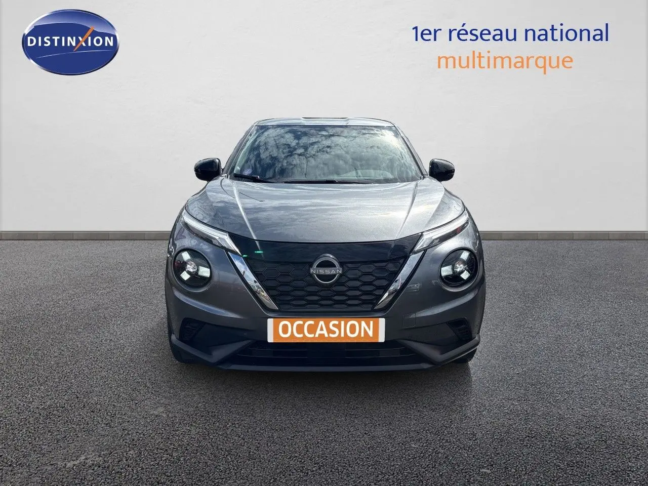 Vue frontale d'un Nissan Juke Hybrid 143ch Business Edition gris squale métal avec calandre noire et phares ronds allumés.