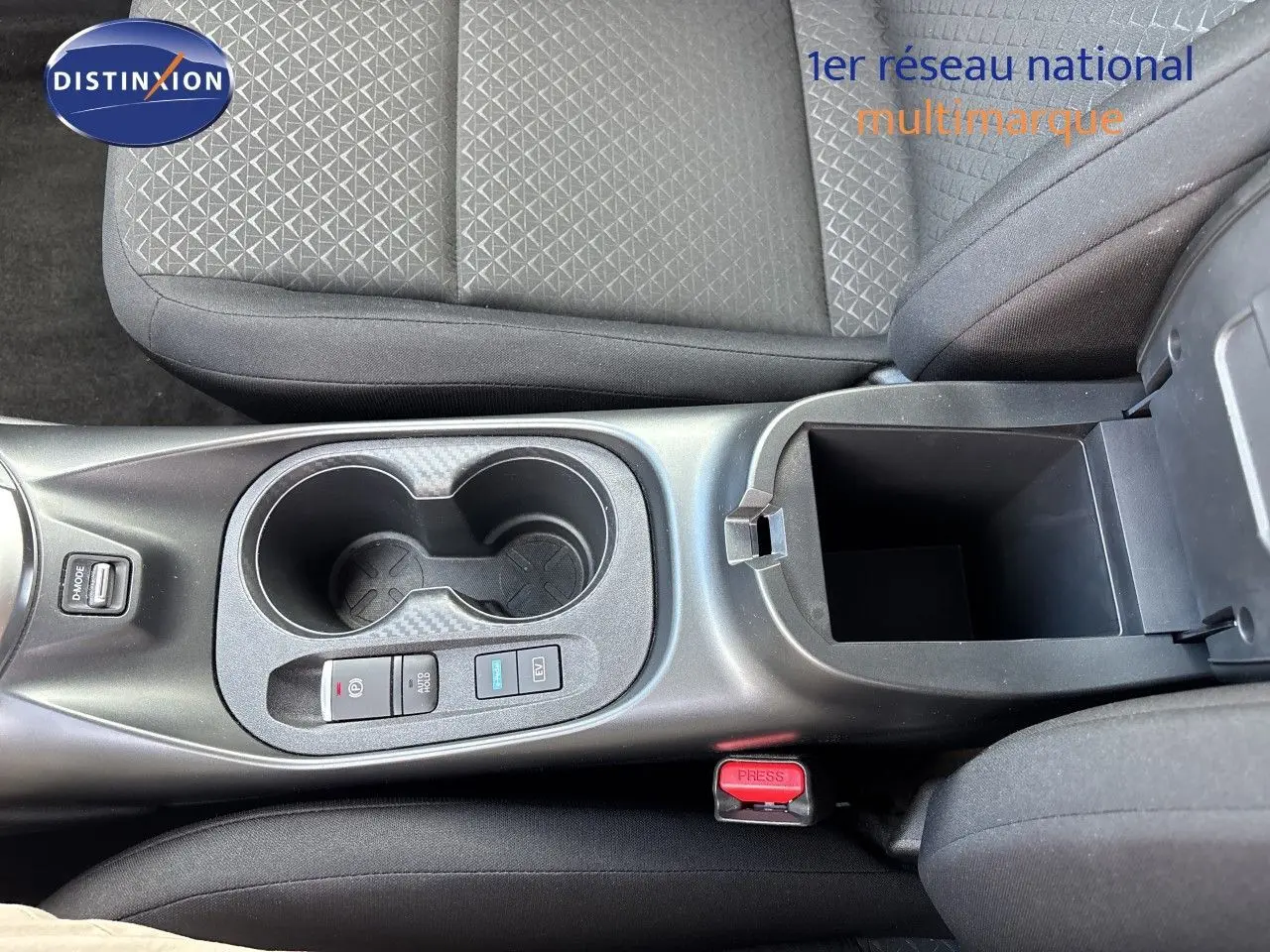 Vue plongeante sur la console centrale du Nissan Juke Hybrid 2024, avec porte-gobelets et rangement ouvert, sièges tissu noir.