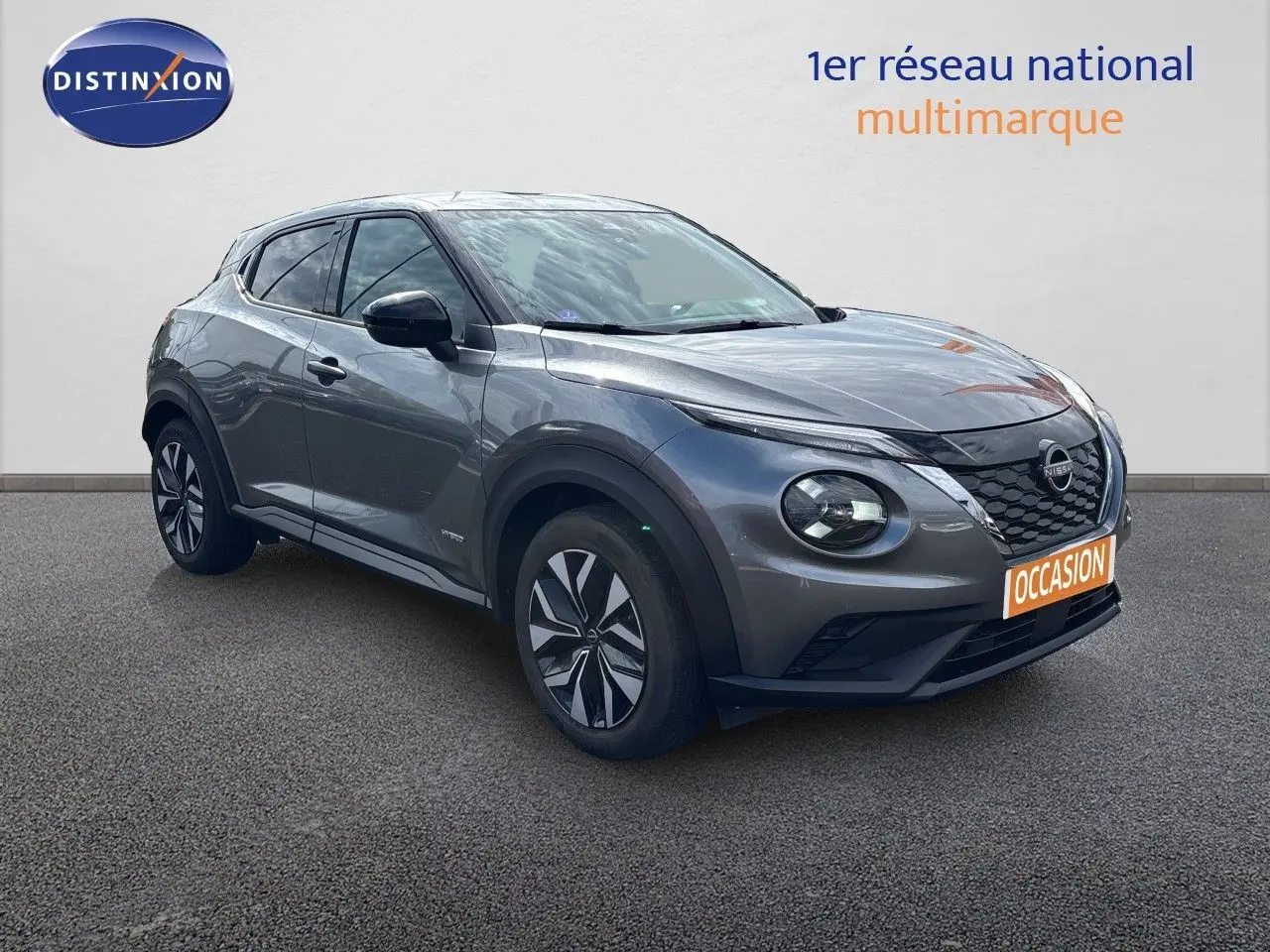 Nissan Juke Hybrid gris squale metal en 3/4 avant droit, avec jantes stylisées et calandre noire distinctive.