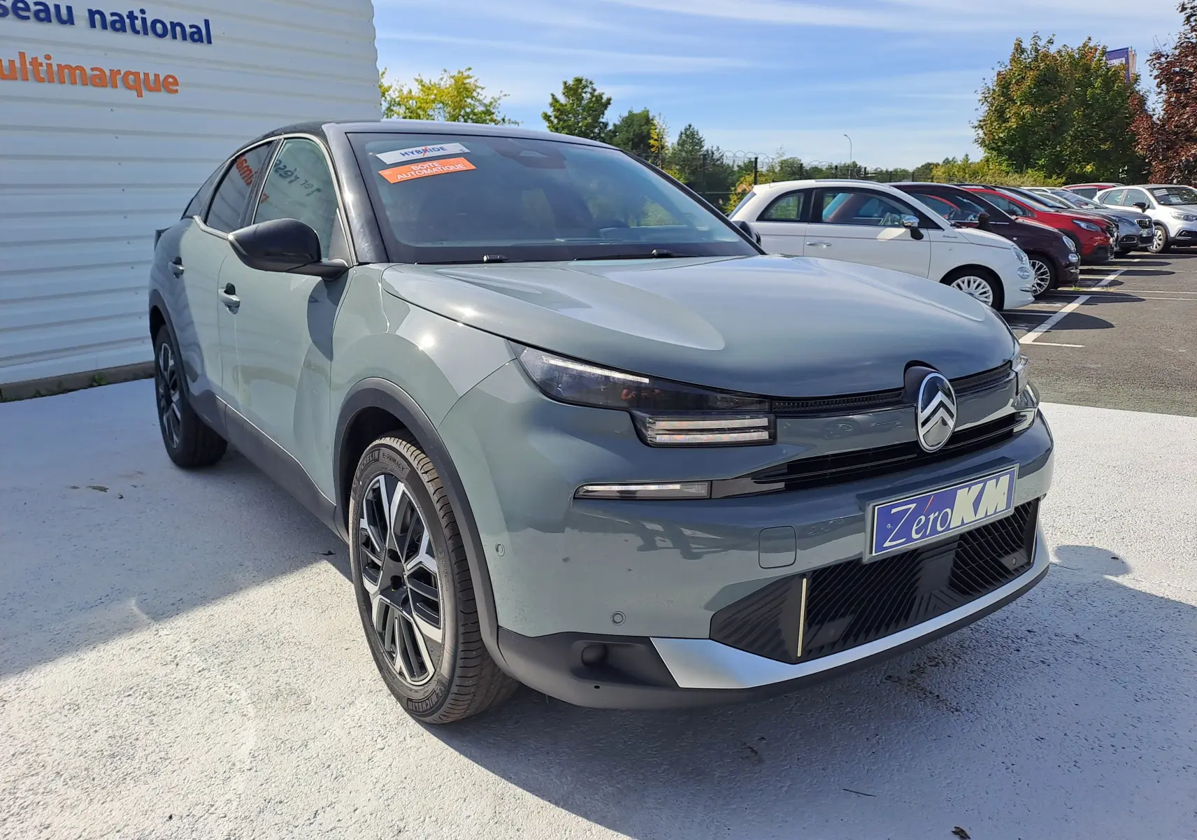 Vue 3/4 avant d'une Citroën C4 hybride 2025 Manhattan Green avec toit noir, sur parking extérieur ensoleillé.