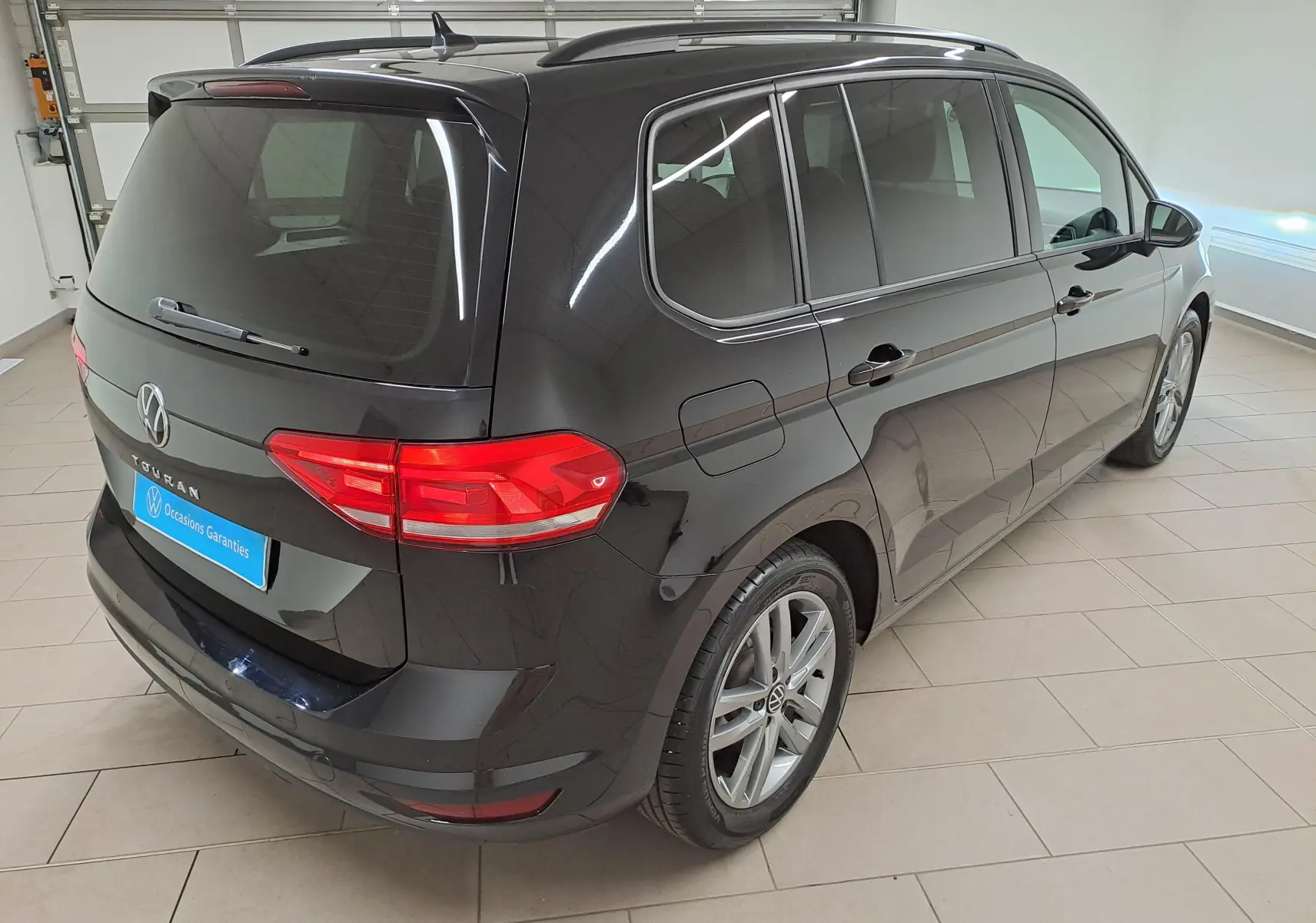 Vue 3/4 arrière droite d'un Volkswagen Touran 2025 noir grenadille avec feux arrière allumés en intérieur.