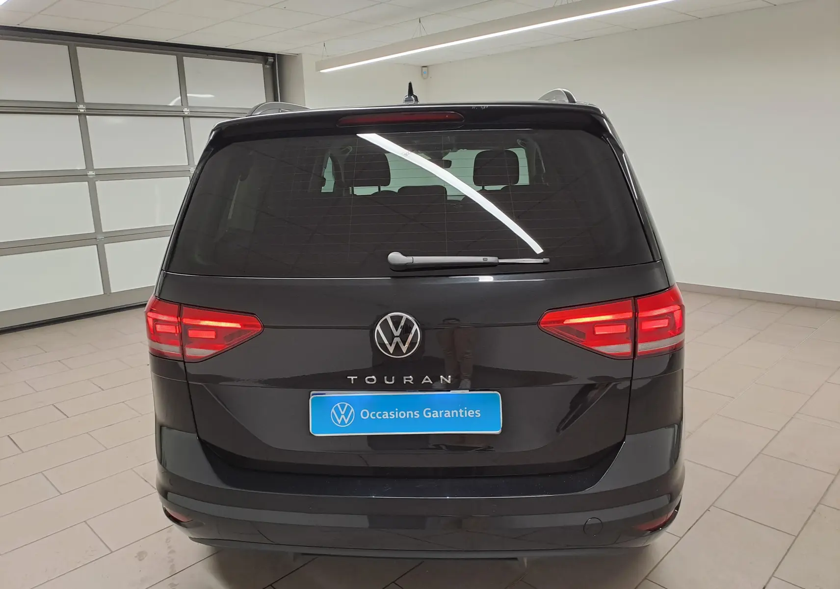 Vue arrière d’un Volkswagen Touran noir grenadille 2025 avec feux allumés dans un garage lumineux.
