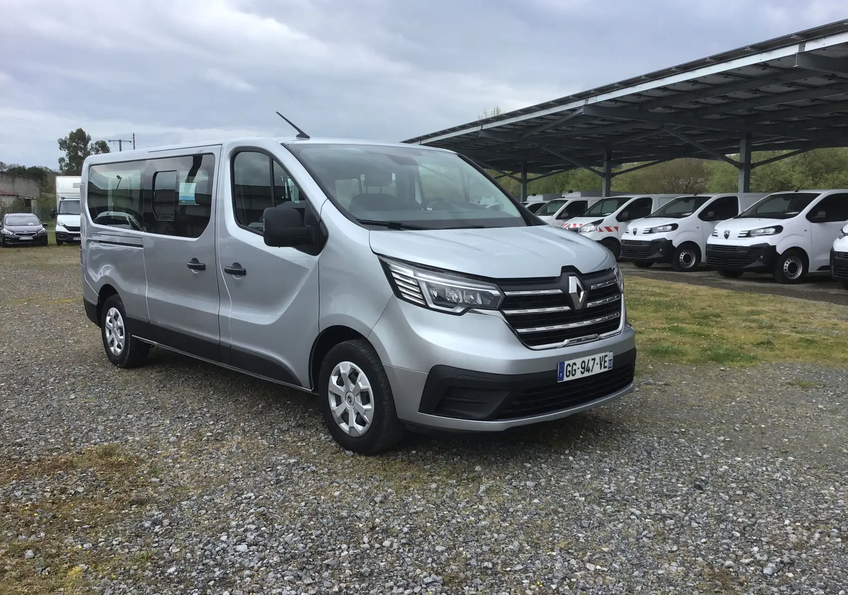 Renault Trafic Combi gris clair métal vu en 3/4 avant droit sur un parking avec plusieurs véhicules blancs en arrière-plan.