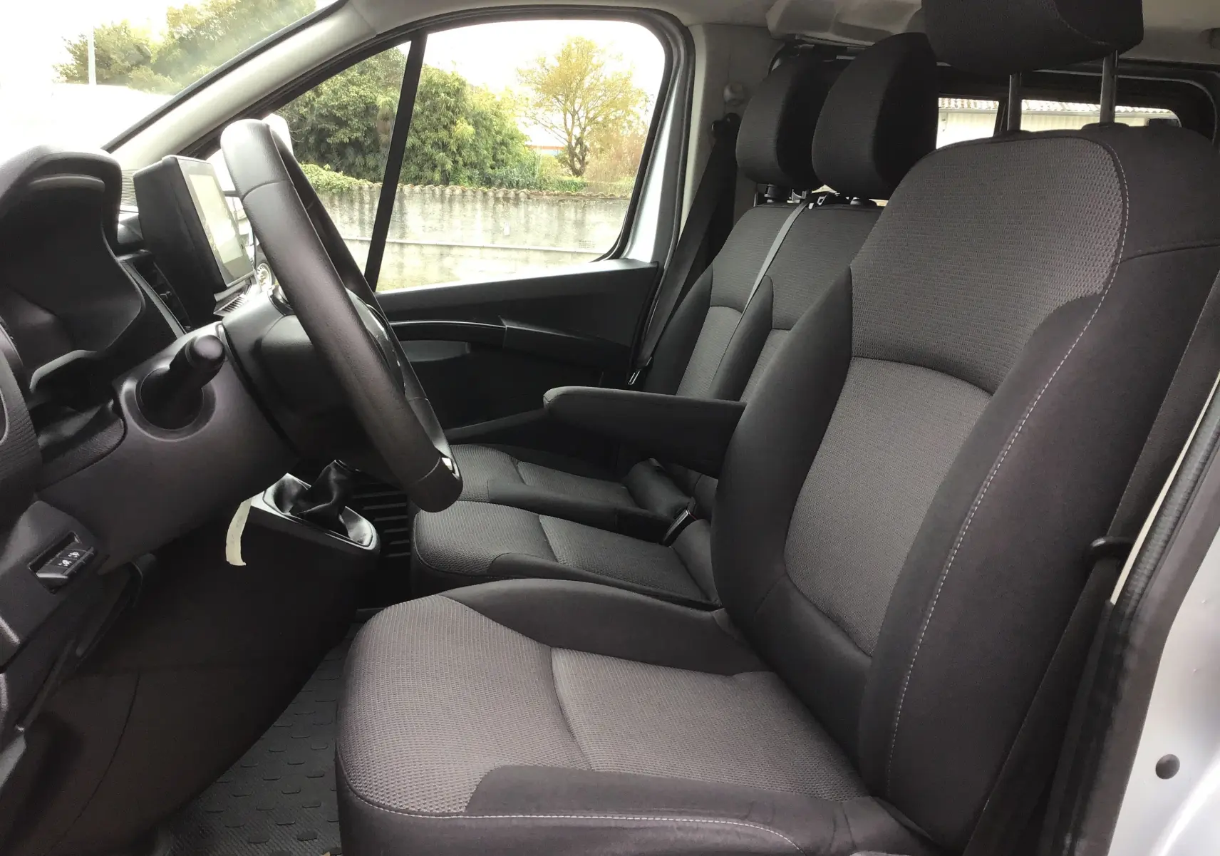 Vue intérieure côté conducteur du Renault Trafic Combi 2022, sièges tissu gris foncé avec accoudoir et volant multifonction.