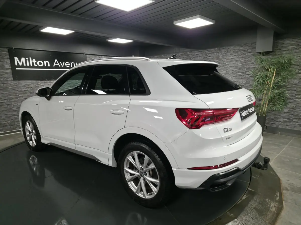 Audi Q3 blanc en 3/4 arrière droit, avec jantes S line et attelage amovible visible, dans un showroom intérieur.