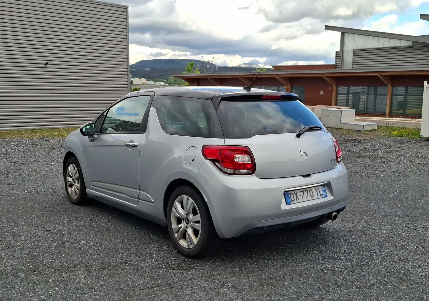 Vue 3/4 arrière droite d'une Citroën DS3 gris clair avec toit noir et vitres arrière surteintées, garée sur un sol gravillonné.