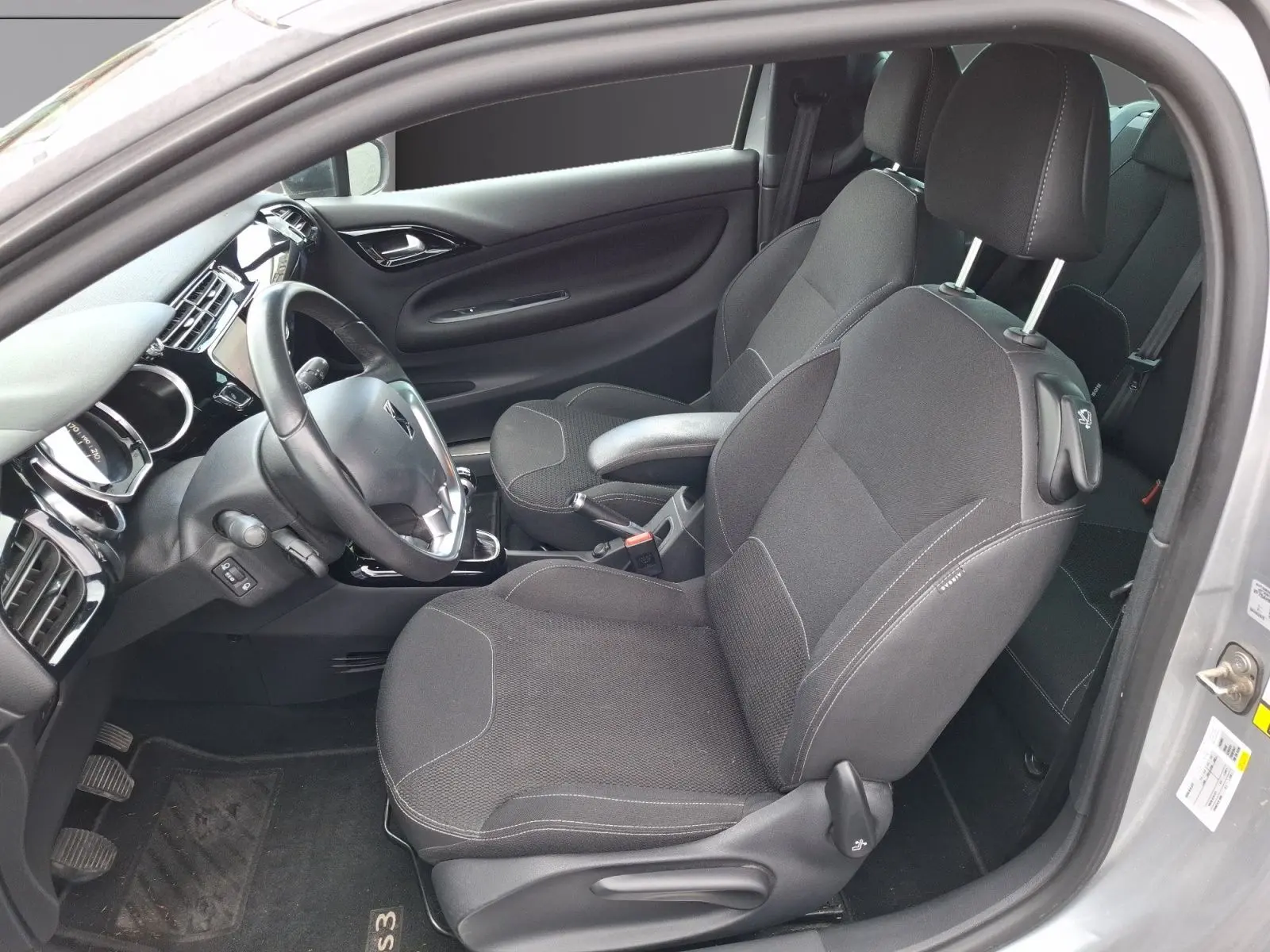 Vue intérieure côté conducteur d'une Citroën DS3 gris clair de 2015, sièges tissu noirs et tableau de bord moderne.