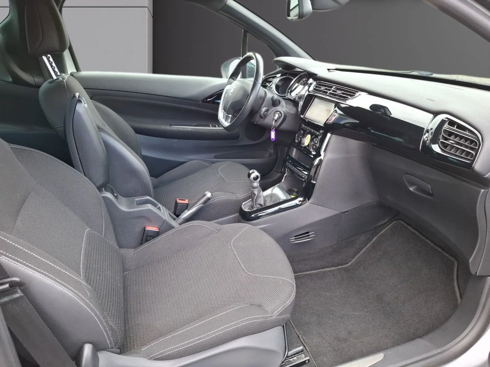 Vue intérieure côté droit de la Citroën DS3 2015 avec sièges gris, tableau de bord noir brillant et boîte manuelle.