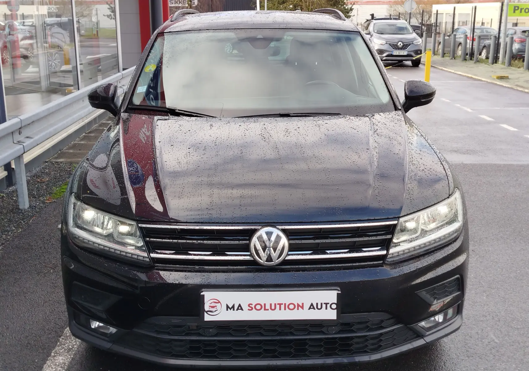 Vue de face d'un Volkswagen Tiguan noir intense nacré 2019 avec phares allumés et gouttes de pluie visibles sur la carrosserie.
