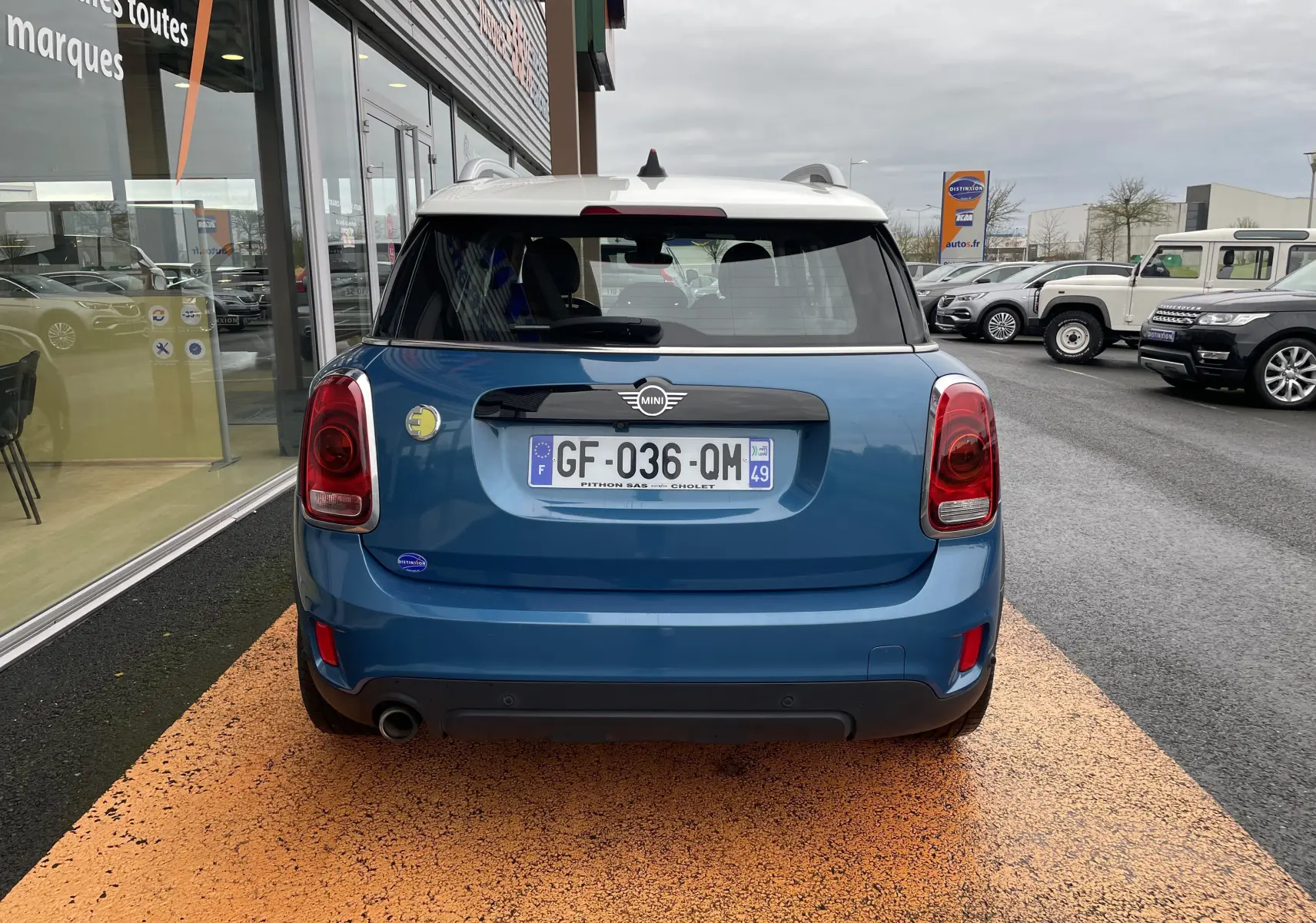 Vue arrière d'une MINI Countryman Cooper S hybride rechargeable bleu Island Metal avec toit blanc, stationnée devant un showroom.